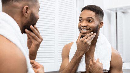 Happy afro man with bare torso touching beard looking in mirror at bathroom. Modern man in bathroom...
