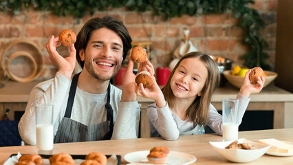 Homemade Bakery. Happy young dad and his cute daughter posing with fresh baked muffins in hands, baking together at home, smiling at camera