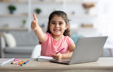 Distant Learning. Little Arab Girl Showing Thumb Up While Study With Laptop At Home, Smiling Female Child Sitting At Table With Computer In Living Room, Recommending Online Education, Copy Space