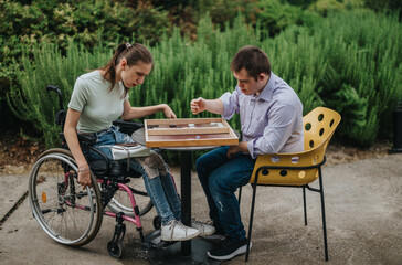 A boy with Down syndrome and a girl in a wheelchair enjoy a game of backgammon together outside. A...