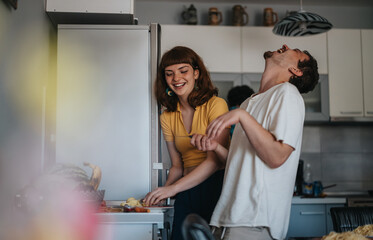 Two young friends share a joyful moment as they laugh together in a modern kitchen setting,...