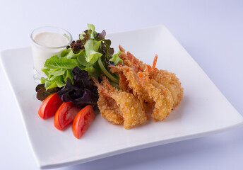 Fried shrimp salad with green lettuce, green oats, red tomatoes and white dressing on a white square plate on a white backdrop.
