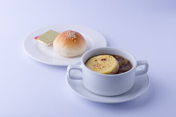Clear onion soup in a white bowl, with sliced ​​potatoes, served with buttered bread on a white plate, on a white backdrop.