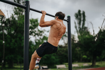 Athletic man doing pull-ups on a metal bar in an outdoor park. Strength training and fitness under...
