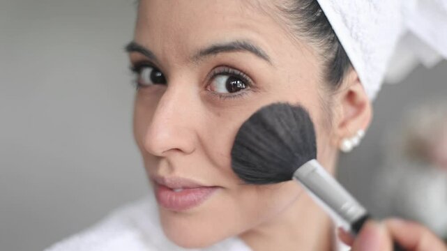 Beautician applying makeup to her cheek with a brush, showcasing a towel wrapped head and a focus on beauty and care