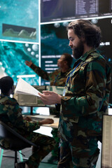 Special operations engineer reviewing operation case file before briefing the squad in military command post, examining crucial information from satellite CCTV surveillance system.