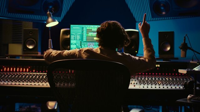 Cheerful energic audio engineer dancing on his own music creation in professional studio, editing a recording on mixing console and daw software. Sound designer processing tracks. Camera B.