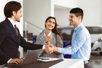 Young Spouses Buying Car, Husband Handshaking With Seller Man Standing In Dealership Office. Selective Focus