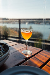 glowing orange cocktail in nick and nora glass on outdoor table overlooking water on sunny day