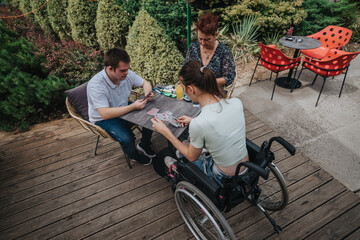 A boy with Down syndrome, a girl in a wheelchair, and an older woman play cards together outside,...