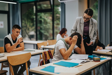 Students in a classroom setting, one looking stressed and receiving guidance from a teacher,...