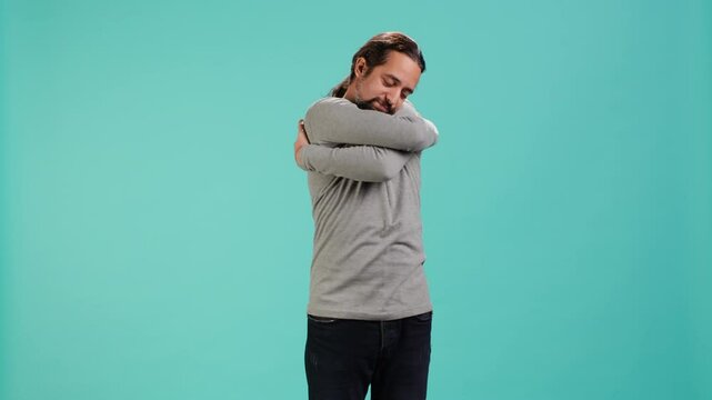 Portrait of man standing in studio, hugging himself in gesture of comfort and self love, isolated over studio background. Cheerful person doing self acceptance, embracing himself, camera A