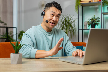 Asian man wearing headset, freelance worker, call center or support service operator helpline, having talk with client or colleague communication support at home room. Guy sits on couch at table desk