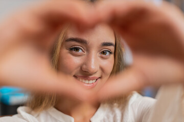 I love you. Portrait of happy Caucasian young woman at home living room couch makes symbol of love, showing heart sign to camera, express romantic feelings express sincere positive feelings. Lifestyle