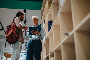 An elderly mentor discusses educational material with a young student in a modern library,...