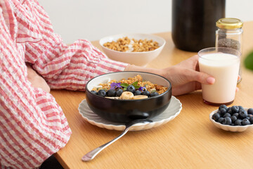 A woman is sitting at a table, eating a healthy breakfast. Granola with Greek yogurt and blueberries