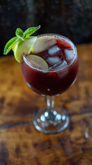 Copa de sangría rojo oscuro con hielo, rodajas de manzana y hojas de menta, sobre mesa de madera...