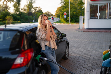 Beautiful blonde woman drinks coffee while gassing up her car, idyllic sunset on gas station 