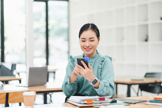 Businesswoman checking an email notification on smartphone, with laptop displaying financial charts at home office.