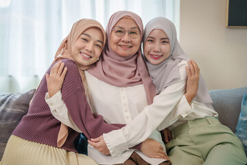 cheerful senior mother and adult daughters in hijab dresses sitting on sofa,embracing each other,posing to camera,family lifestyle,meeting and relationship