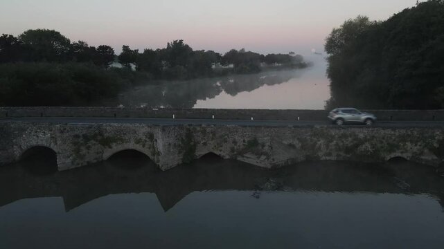 A van and car cross a stone bridge over calm water at dawn in Adare town Ireland