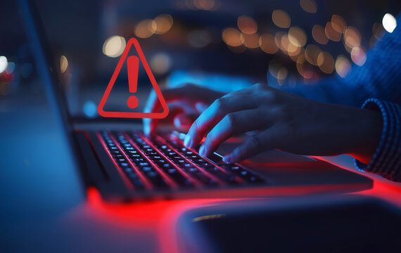 A person types on a laptop, illuminated in red, with a warning symbol highlighted, indicating a potential cybersecurity issue.