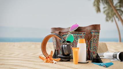 Beach essentials set on sandy shore: a stylish bag with sunglasses, a hat, headphones, sunscreen,...