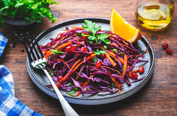 Vegan coleslaw salad with red cabbage, carrot, parsley, pomegranate, orange and olive oil dressing on wood background, top view