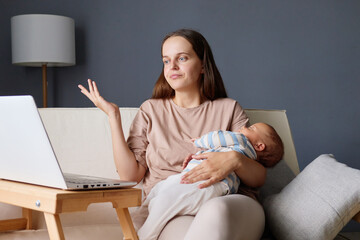 Puzzled woman sitting on couch with her baby in hands talking on video call shrugging unsure about deadline balancing business tasks and motherhood from her home workspace during maternity leave