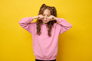 Sad upset schoolgirl with curly hair wearing fashionable sweatshirt crying expressing sadness being offended wiping tears with her fists rubbing eyes isolated over yellow background