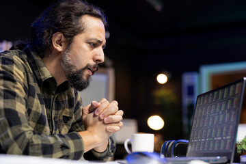 Young guy seated at his desk, carefully examining stock data on his laptop. Freelancer, a man, focused intently on forex trading chart on his device while examining the economic trend value.