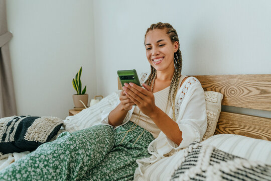 Happy woman sitting on bed and using smart phone at home