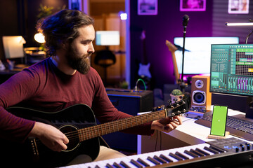 Audio engineer performing on guitar in his home studio, watching video tutorials and practicing on strings. Musician learning multiple songs on acoustic instrument, uses smartphone greenscreen.