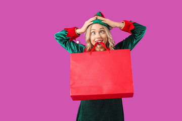 Funny young woman dressed as elf with shopping bag on purple background