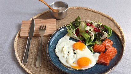 Bright sunny eggs with orange yolks beside green salad, pomegranate seeds, and salmon slices on blue plate, served with silver bowl of salt, emphasizing eco-conscious healthy lifestyle.