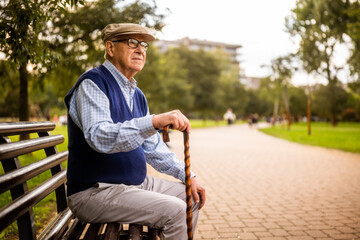 Portrait of worried senior man in park. He is sitting on bench on autumn day.