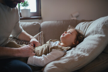 Father taking care of sick girl with fever and cold at home. Daughter lying on sofa, feeling tired.