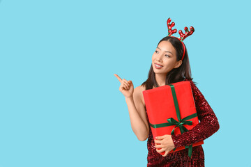 Young Asian woman in reindeer horns holding Christmas gift box and pointing at something on blue...