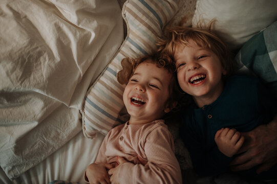 Candid portrait of siblings lying together in bed, looking at the camera and laughing. Top view.