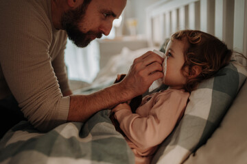 Sick girl with cold lying in the bed. Father helping little daughter to blow her nose.