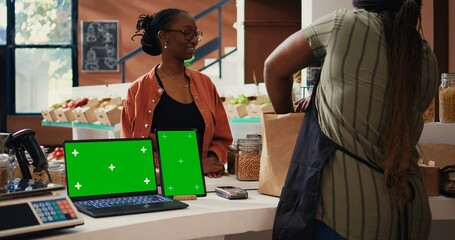 Client buying bio produce next to greenscreen at cash register, supporting farming small business. Storekeeper using blank copyspace display on laptop and tablet placed on counter. Tripod shot.