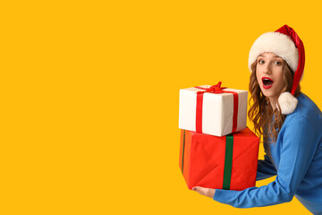 Surprised young woman in Santa hat with Christmas gift boxes on yellow background