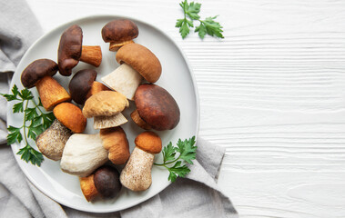 A variety of fresh mushrooms displayed on a plate surrounded by green herbs on a simple wooden surface