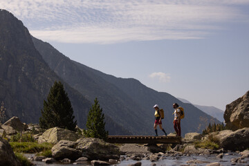 Fototapeta premium Two people are walking across a bridge over a river. The mountains in the background create a sense of awe and wonder. The scene is peaceful and serene, with the sound of the water