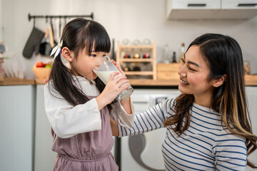 Asian little daughter drinking a glass of milk with mother in kitchen. 