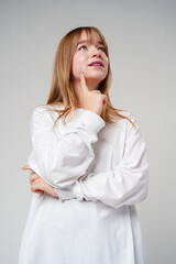 Young woman in a white long-sleeve shirt thoughtfully poses with a hand on her chin in a minimalistic setting