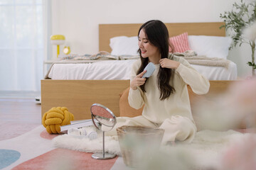 Asian woman doing self hair care in cozy bedroom, Young female is doing her hair with a comb and smiling while looking at the mirror in the bedroom at home. Lifestyle and daily activities