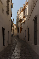 narrow, cobbled street in a white village, with houses on both sides and the sunlight at dawn reflecting in the window of one of them.