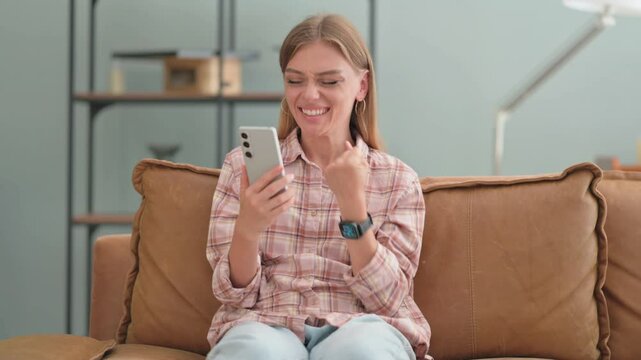 Young Woman Excited for Results on Phone while Sitting on Sofa at Home