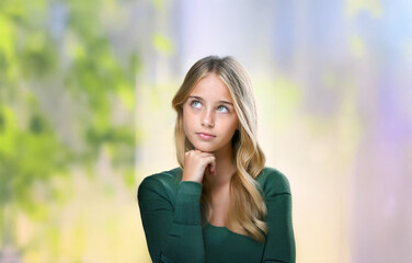 A young woman with long blonde hair and green eyes is looking up and pondering something with her hand on her chin.